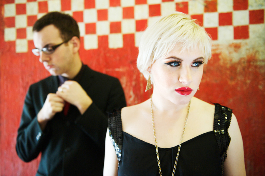 Man and woman standing in front of a red checkered wall, with solemn expressions