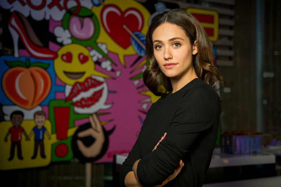 Actress Emmy Rossum stands in front of a colorful wall, with a soft expression