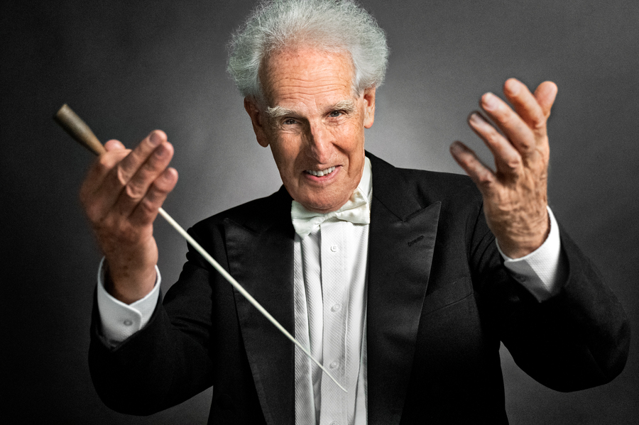 Conductor Ben Zander standing in front of a gray background, with his hands up and an inviting smile