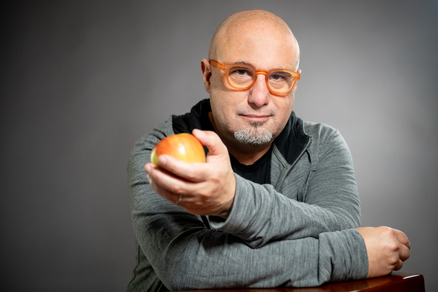 Man with colorful glasses holding an apple up to the camera, with an inquisitive expression