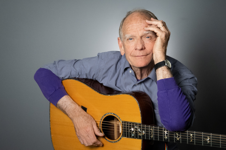 Musician sitting in front of a gray background holding a guitar, with calm demeanor